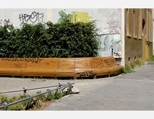 Eine geschwungene Holzbank mit Graffiti steht vor einer Wand, die ebenfalls mit Graffiti bedeckt ist. Im Hintergrund sind Büsche und ein gepflasterter Bürgersteig zu sehen, daneben befindet sich ein gelblich-braunes Gebäude.