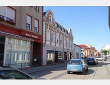 Straßenszene in einer kleinen Stadt mit mehrstöckigen, historischen Gebäuden auf der linken Seite, darunter ein Haus mit der Aufschrift "Willy Ernst". Im Vordergrund stehen parkende Autos, und im Hintergrund sind weitere Gebäude und ein klarer Himmel zu sehen.