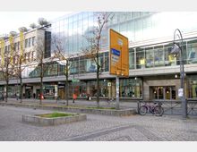 Straßenszene mit einem modernen Gebäude mit Glasfassade, Geschäften im Erdgeschoss und einer großen gelben Wegweiser-Tafel im Vordergrund. Im Straßenraum sind Bäume, Fahrräder und Fußgänger sowie gepflasterte Flächen sichtbar.