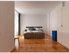 Schlafzimmer mit minimalistischem Design, einer zentral platzierten Doppelbett mit neutraler Bettwäsche, einem hölzernen Boden, weiß gestrichenen Wänden, großen Fenstern mit Vorhängen und einem weißen Kleiderschrank rechts. Im linken Vordergrund steht eine Holzbank.