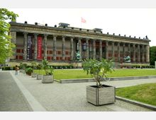 Außenansicht des Alten Museums in Berlin, einem klassizistischen Gebäude mit einer Säulenfassade und lateinischer Inschrift. Im Vordergrund ein gepflasterter Weg, grüne Rasenflächen sowie Pflanzkästen; rechts steht eine Bronzestatue.