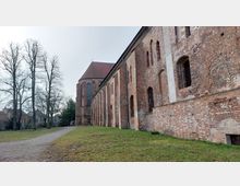 Backsteinbau einer historischen Kirche mit hohen gotischen Fenstern und einem spitz zulaufenden Dach, umgeben von einer Wiese und kahlen Bäumen in einer ländlichen Umgebung.