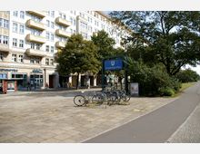 Ein U-Bahn-Eingang mit blauem Schild "Strausberger Platz" steht vor einem mehrstöckigen Wohngebäude mit Balkonen und Geschäften im Erdgeschoss. Im Vordergrund sind Fahrradständer mit mehreren angelehnten Fahrrädern, daneben verläuft ein gepflasterter Platz mit Bäumen.