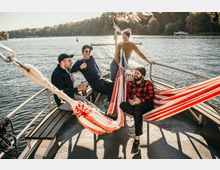 Vier Personen entspannen auf einem kleinen Boot auf einem See, darunter zwei in rot-weiß-gestreiften Hängematten. Umgeben von ruhigem Wasser und Bäumen im Hintergrund, sind auf dem Boot Holzbänke, ein Metallgeländer und bunte Wimpel zu erkennen.