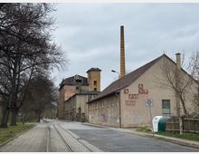 Straßenszene mit einem alten Industriegebäude, das teilweise renoviert wurde, und einer angrenzenden Sporthalle mit einem gemalten Schriftzug „Sporthalle“ an der Fassade. Im Vordergrund verläuft eine gepflasterte Straße mit Schienen, rechts steht ein Verkehrsschild für eine 30er-Zone, links sind kahle Bäume zu sehen.