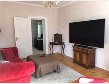 Wohnzimmer mit Holzboden und heller Wandfarbe, ausgestattet mit einem roten Sofa, einem kleinen Tisch mit brauner Tischdecke, einem großen Fernseher auf einem antiken Holzschrank und einem kleinen Beistelltisch. Im Hintergrund ist eine offene Tür, die in ein Arbeitszimmer mit Schreibtisch und Regalen führt.