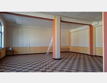 Leerer Innenraum eines größeren Raums mit kariertem Fliesenboden, abgehängter Rasterdecke und mehreren tragenden Säulen in Creme- und Rottönen. An der Wand stehen eine Leiter sowie Heizkörper und ein kleines Tischchen, links sind hohe Fenster zu sehen.