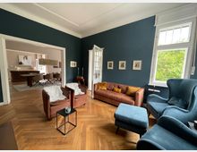 Wohnzimmer mit dunkelblauen Wänden, Parkettboden und großen Fenstern, die Tageslicht hereinlassen. Möbliert mit einem braunen Ledersofa, zwei blauen Sesseln, einem kleinen schwarzen Beistelltisch und mehreren dekorativen Bildern an der Wand; im Hintergrund ein Essbereich mit Esstisch und Stühlen.