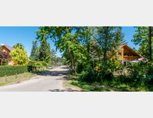 Ruhige Wohnstraße in einer ländlichen Umgebung mit freistehenden Einfamilienhäusern, dichtem Baumbestand und gepflegten Gärten. Ein Haus mit holzverkleideter Fassade und Balkon steht rechts im Bild, während links Hecken und Bäume entlang der Straße verlaufen.