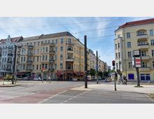 Straßenkreuzung in einer Stadt mit mehrgeschossigen Altbaugebäuden, gelber Fassade und kleinen Balkonen; im Erdgeschoss befinden sich Geschäfte und ein Restaurant mit rotem Markisen-Dach. Eine Straßenbahnlinie verläuft durch die Kreuzung, und Fußgängerampeln sowie eine Uhr auf einem Mast sind sichtbar.