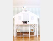 Ein Kinderzimmer mit einer Hochbettkonstruktion in Form eines kleinen weißen Häuschens, verziert mit Sternen. Darunter befinden sich Aufbewahrungskörbe und Spielzeug auf einem Holzregal, während eine getigerte Katze auf der Kante des Bettes sitzt; der Raum hat eine mintgrüne Rautentapete.
