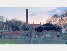 Ruine eines historischen Backstein- und Steingebäudes mit teilweise eingestürztem Dach, sichtbarem Schornstein und zerfallenen Fassaden. Im Hintergrund sind kahle Bäume und ein Sonnenuntergang zu sehen, während ein Zaun das Gelände umgibt.