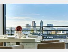 Blick von einem modern eingerichteten Balkon auf eine städtische Skyline mit hohen Bürogebäuden. Im Vordergrund steht ein weißer Tisch mit Geschirr und einer Vase mit roten Blumen, umgeben von beigen Stühlen.