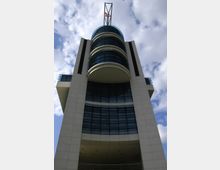 Futuristisches Hochhaus mit einer hellen Steinfassade und mehreren gebogenen Glasfronten, gekrönt von einer Kuppel und einer filigranen Stahlstruktur, vor einem bewölkten Himmel.