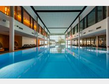 Großer Innenpool in einem modernen Wellnessbereich mit klarer blauer Wasseroberfläche, flankiert von Liegestühlen auf beiden Seiten. Die Architektur umfasst hohe Fensterfronten und weiße Säulen, die viel Licht in den Raum lassen.