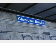 Blaues Schild mit weißer Aufschrift „Glienicker Brücke“ an einer grauen Steinmauer unter einer Überdachung mit sichtbaren Metallträgern.