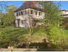 Zweistöckiges Einfamilienhaus mit weißer Fassade und rotem Ziegeldach, umgeben von einem Garten mit Bäumen, einem Rasen und einem kleinen Teich im Vordergrund. Im Hintergrund ist eine teilweise sichtbare Nachbarbebauung erkennbar.