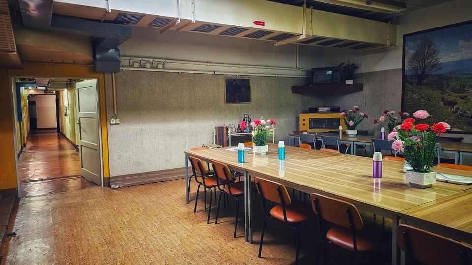 Innenraum eines schlichten Aufenthalts- oder Speiseraums mit langen Holztischen und Stühlen, auf den Tischen stehen Vasen mit Blumen und Thermoskannen. An der Decke verlaufen sichtbare Lüftungskanäle und Leuchtstoffröhren; rechts hängt ein großes Landschaftsbild, links führt eine offene Tür in einen langen Flur.