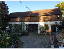 Zweistöckiges Gebäude mit weißer Fassade im unteren Bereich und unverputzten Ziegeln im oberen Bereich, umgeben von einem gepflasterten Hof mit Pflanzen und Bäumen. Im Vordergrund sind Sitzbereiche, Holztreppen und Blumenkübel zu sehen, während das Gebäude große Holztüren und halbkreisförmige Fenster besitzt.