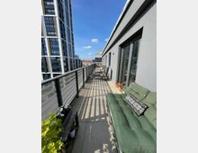 Langer Balkon einer modernen Wohnung mit grauem Boden und einer Glas- und Metallgeländerbegrenzung. Darauf stehen Sitzmöbel, darunter ein grünes Sofa und ein Tisch mit Pflanzen. Im Hintergrund sind ein hohes Wohngebäude und ein klarer blauer Himmel sichtbar.