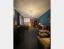 Innenraum eines Cafés oder einer Lounge mit hohen Decken, Kronleuchter und farbigen Wänden in Blau und Bordeaux über freiliegendem Ziegel. Mehrere Tische und Stühle, Sofas, Stehlampen, ein Regal mit Büchern sowie ein Schachbrettbodenbereich und eine Leinwand an der Rückwand sind zu sehen.