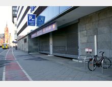 Außenansicht eines Parkhauses mit grauer Steinfassade und verschlossenem Rolltor, darüber Beschilderung mit „Einfahrt“ und „Ausfahrt.“ Im Vordergrund führt ein Radweg vorbei, ein Fahrrad steht an einem Metallständer, und im Hintergrund ist ein historisches Gebäude mit Turm erkennbar.