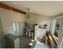 Modernes Badezimmer mit hellen Wänden, freiliegendem Holzbalken und großen Fenstern, die Tageslicht hereinlassen. Zu sehen sind eine freistehende Badewanne, ein Waschtisch mit Waschbecken, eine bodengleiche Dusche mit hellgrünen Fliesen und ein WC; natürliche Dekorationselemente wie Trockenblumen und Holzakzente ergänzen den Raum.