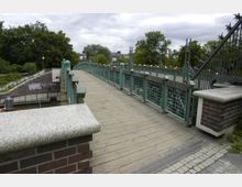 Eine Fußgängerbrücke mit grünem Geländer und Holzplanken führt über einen Kanal. Umgeben von Bäumen und Büschen ist im Hintergrund ein grauer Himmel und einzelne Gebäude sichtbar, während die Brücke durch Backsteinpfeiler abgestützt wird.