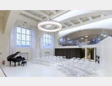 Moderner Veranstaltungssaal mit hohen Decken, großen Bogenfenstern und einer hellen, minimalistischen Einrichtung. Im Vordergrund stehen ein schwarzer Flügel und weiße Stühle, während im Hintergrund eine Galerieebene mit Glasgeländer und eine Bühne mit Vorhang sichtbar sind.