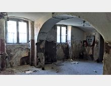 Ein heruntergekommener Raum mit verputzten Wänden, von denen der Putz abblättert, zwei Fenstern mit dünnen Vorhängen und einem Gewölbebogen an der Decke. Der Boden ist uneben und von Schmutz bedeckt, mit einer Tafel oder einem Plakat an einer der beschädigten Wände.