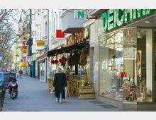 Straßenszene mit Geschäftsfassaden, darunter ein asiatisches Restaurant mit roten Lampions und Sitzgelegenheiten sowie ein Schuhladen mit großen Schaufenstern. Auf dem Gehweg sind Passanten, ein Motorroller und Straßenschilder zu sehen.