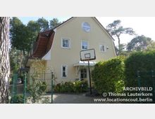 Zweistöckiges Einfamilienhaus mit gelber Fassade, roten Dachziegeln und weißen Fenstern. Vor dem Haus stehen ein Basketballkorb, Sträucher und ein Zaun, umgeben von Bäumen in einer bewaldeten Umgebung.