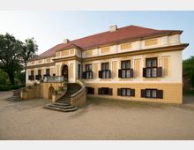 Großes Herrenhaus im barocken Stil mit gelben und weißen Fassadenelementen, symmetrischen Fenstern mit braunen Fensterläden und einem roten Walmdach. Vor dem Eingang eine breite, geschwungene Freitreppe, umgeben von Gartenflächen.