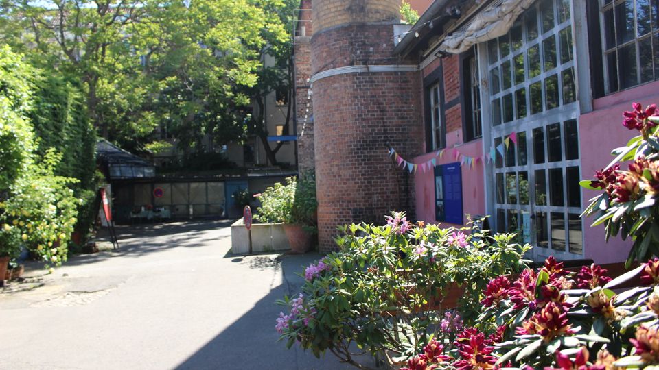 Innenhof mit Backsteinbau und rundem Turm; rechts eine Fassade mit großen Fensterfeldern und blauem Eingang, dekoriert mit Wimpelgirlanden. Im Vordergrund stehen blühende Sträucher und Kübelpflanzen, links säumen Bäume und Grün die asphaltierte Zufahrt.