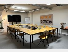 Moderner Besprechungsraum mit U-förmig angeordneten Holztischen und Holzstühlen auf grauem Teppichboden. An der Wand hängen ein großes Display, ein Whiteboard und ein Plakat mit der Aufschrift „MEET ME AT DISTRICT ONE“, daneben eine Pflanze und ein kleiner Tisch mit Dekoration.