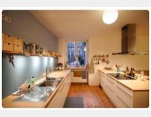 Moderne Küche mit hellem Holzfußboden, weißen Schränken und einer Arbeitsplatte auf beiden Seiten. Ein großes Fenster in der Mitte lässt Tageslicht herein, während an der linken Wand Regale mit Küchenutensilien angebracht sind. Eine Edelstahlspüle und ein Kochfeld mit Dunstabzugshaube sind sichtbar.