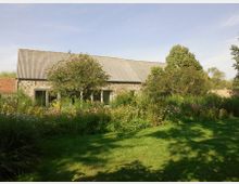 Steinhaus mit Wellblechdach in einer ländlichen Umgebung, umgeben von Bäumen, Blumenbeeten und einer Rasenfläche im Vordergrund. Der Himmel ist klar und sonnig.