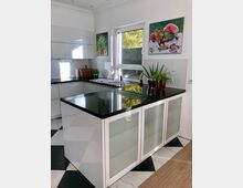 Moderne Küche mit weißer Einbauküche, schwarzer Arbeitsplatte und einer zentralen Kücheninsel mit Milchglasfronten. Ein großes Fenster lässt Tageslicht herein, und an den Wänden hängen zwei farbenfrohe Stillleben-Gemälde; Topfpflanzen und Küchenutensilien befinden sich auf den Arbeitsflächen.