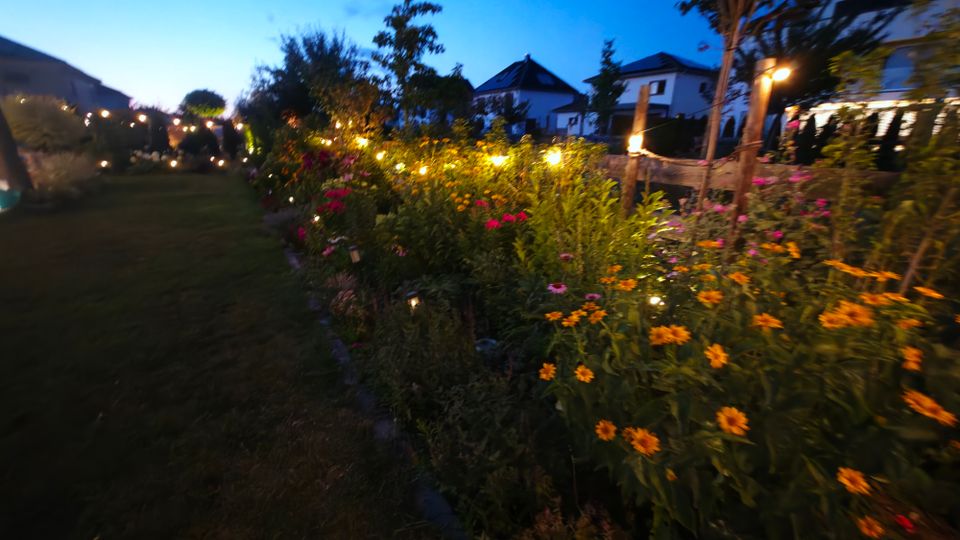 Beleuchteter Garten in der Dämmerung mit breiten Blumenbeeten und einem Rasenweg, gesäumt von Lichterketten und kleinen Bodenleuchten. Im Hintergrund stehen mehrere Wohnhäuser mit Satteldächern sowie Bäume und Sträucher.