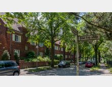 Straßenszene in einem Wohngebiet mit Backsteinhäusern, gesäumt von hohen, belaubten Bäumen. Am Straßenrand stehen parkende Autos, und ein Straßenschild mit der Aufschrift „Eitel-Fritz-Straße” ist im Vordergrund zu sehen.