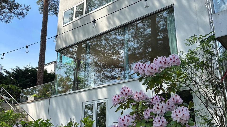 Mehrstöckiges, modernes Wohnhaus mit weißer Fassade, großen Glasfronten und schmalen Fensterbändern, umgeben von hohen Kiefern. Im Vordergrund liegen ein Holzdeck und blühende Sträucher; eine Lichterkette verläuft entlang der Fassade.