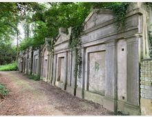 Reihe von historischen Grabmälern aus Stein mit eingewachsenen Efeuranken, gelegen in einem bewaldeten Bereich mit schattigen Bäumen und einem schmalen Erdweg davor. Die Fassaden der Grabmäler zeichnen sich durch Säulen und Dreiecksgiebel aus.
