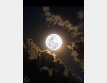 Vollmond am Nachthimmel, umgeben von dunklen Wolken, die teils vom Mondlicht erhellt sind. Sterne sind dezent im Hintergrund sichtbar.