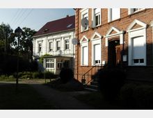 Zwei Wohnhäuser nebeneinander, eines mit roter Backsteinfassade und weißen Fensterrahmen, das andere mit weiß verputzter Fassade und klassizistischen Verzierungen. Im Vordergrund führt ein gepflasterter Weg durch einen kleinen Gartenbereich, umgeben von Büschen, mit einer Straßenlaterne auf der linken Seite.