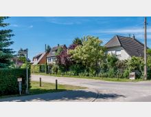 Wohngebiet mit Einfamilienhäusern, umgeben von Bäumen und Hecken. Eine unbefestigte Straße führt durch die Szenerie, neben der ein Straßenschild mit der Aufschrift „Veltener Str.“ steht.