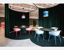 Moderner Innenraum mit zwei runden Tischen, umgeben von farbigen Stühlen (rosa und blau) auf einem dunklen Teppich. Der Bereich ist durch dunkelgrüne Vorhänge abgetrennt, mit farbigen Pendelleuchten und sichtbaren Deckenträgern im industriellen Stil.