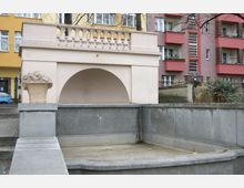 Dekorative, leere Brunnenanlage aus Beton mit einem Steinrelief in Form eines Obstkorbs, umgeben von Wohngebäuden mit bunten Fassaden in einem städtischen Umfeld. Eine Balustrade im Hintergrund über einem bogenförmigen Element ergänzt die Architektur.