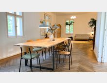 Helles Zimmer mit einem langen Holztisch und mehreren Stühlen in der Mitte, flankiert von großen Fenstern, die Tageslicht hereinlassen. Im Hintergrund befinden sich ein beige Sofa, Zimmerpflanzen und hölzerne Möbel, auf einem Fischgrät-Holzboden.