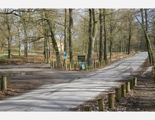Waldweg mit Bäumen und Holzpfosten entlang der Straße, daneben ein Schild mit der Aufschrift „Blockhaus Nikolskoe“. Im Hintergrund ist ein kleines Gebäude zu erkennen.