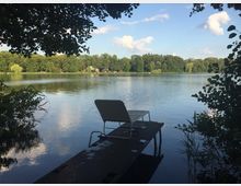 Ein See mit ruhiger Wasseroberfläche, umgeben von dichtem Baumbewuchs im Hintergrund. Im Vordergrund ragt ein kleiner Steg ins Wasser, auf dem ein einzelner Liegestuhl steht, eingerahmt von überhängenden Ästen und Schilf.